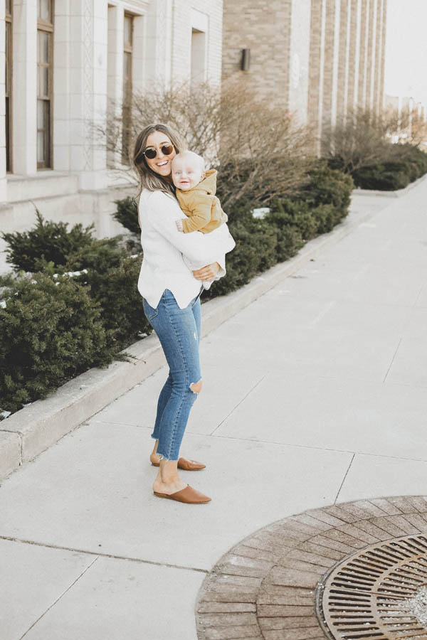 Girl holding her baby in a white sweater and jeans with flats on the sidewalk.